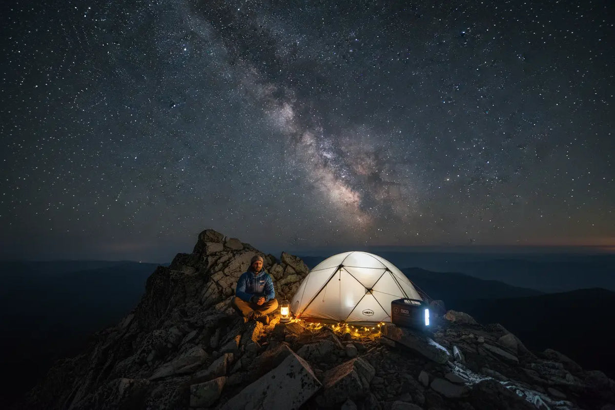 High-altitude camping at night with a portable solar generator powering a glowing tent and lantern under the Milky Way. A mountain climber rests on a rocky summit, using the rugged power bank for essential electronics in freezing outdoor conditions